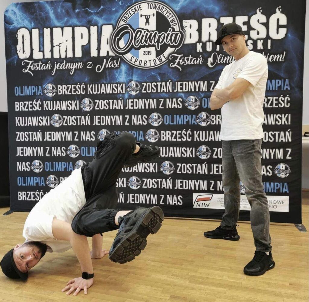 instruktor robi pokaz taneczny  przed banerem zachęcającym do dołączenia do Olimpia Brześć Kujawski 